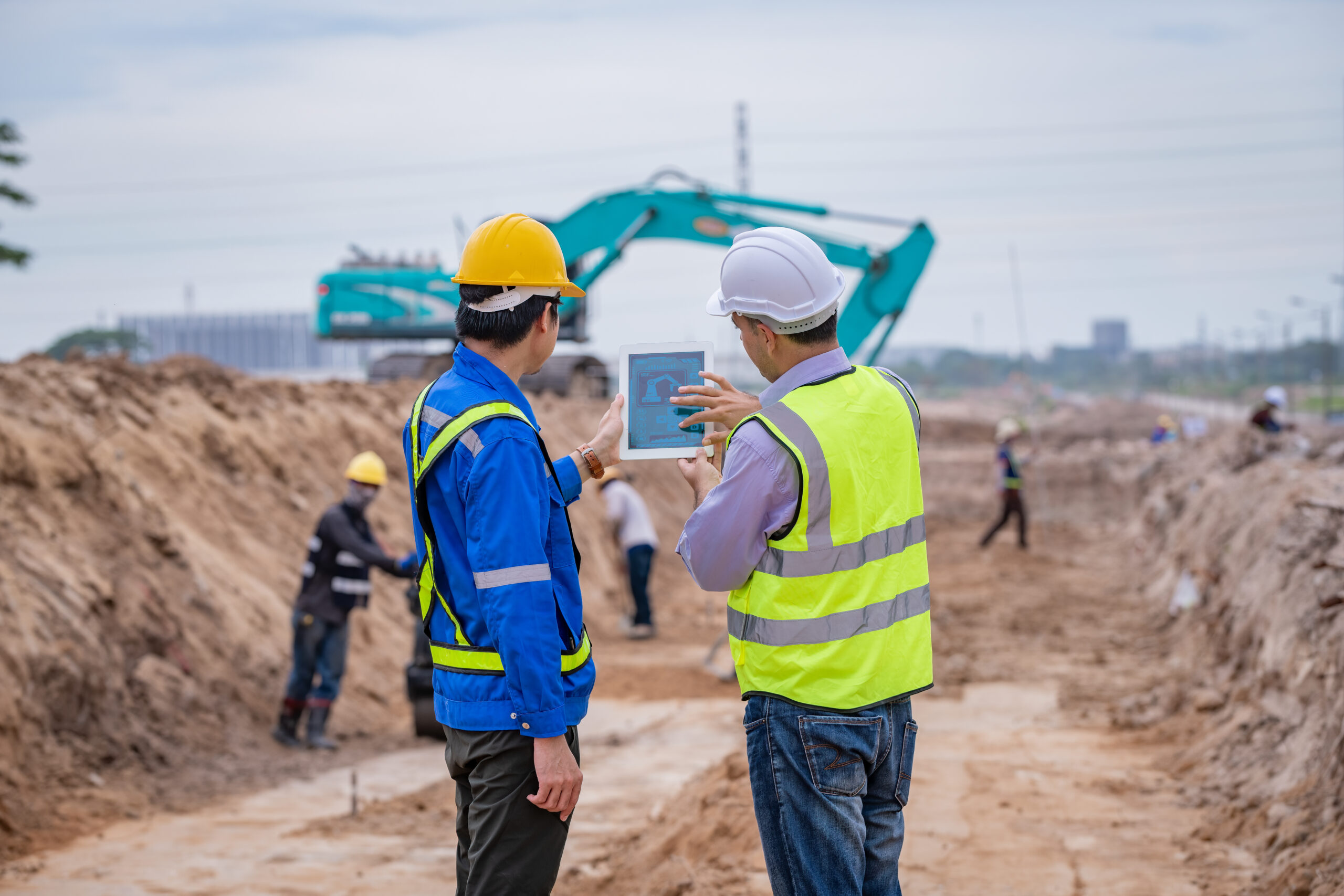 Un vérificateur effectue un examen d'adéquation sur un chantier de construction avec l'utilisateur de la pelleteuse en arrière plan.