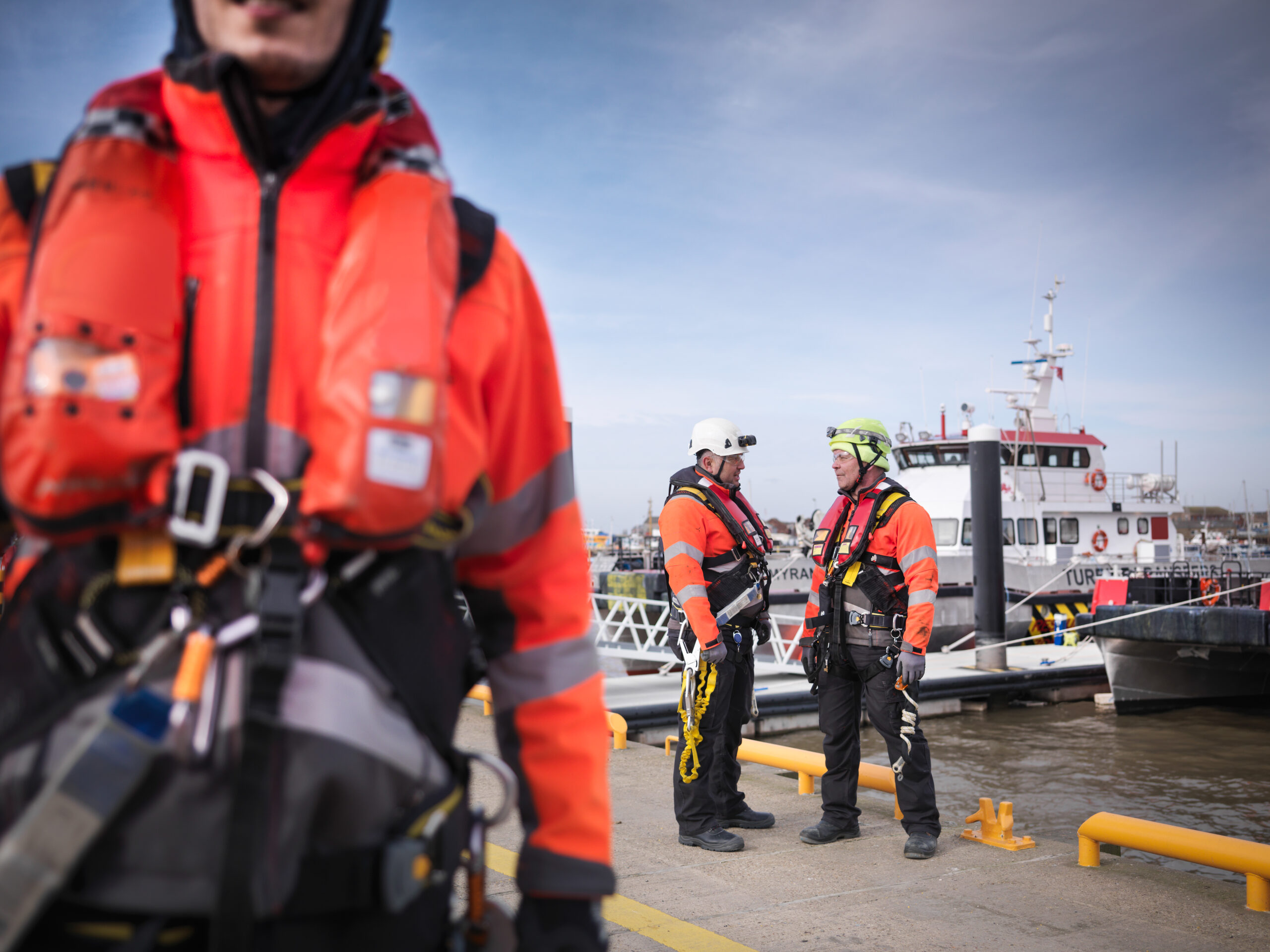 Trois professionnels équipés de gilets de sauvetage travaillent sur un quai portuaire près d’un navire.